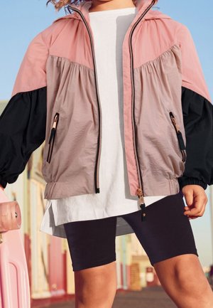 Veste légère bicolore comprenant des sections rose, gris et noir, avec fermeture éclair et poignets élastiques. Associée à un t-shirt blanc et un short noir.