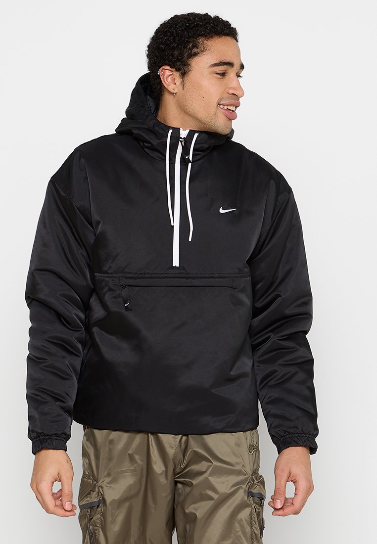 Svart nylon pullover-jacka med huva, dragsko, framficka med dragkedja och elastiska ärmslut. Har en liten vit logotyp på bröstet.