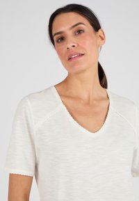 Camiseta blanca de cuello en V con mangas cortas, con un dobladillo decorativo en el cuello y las mangas, hecha de una tela texturizada y ligera.