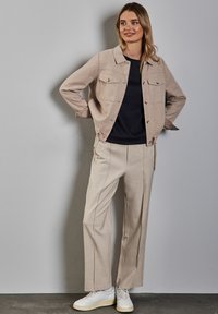 Beige Wollsakko aus einer Wollmischung mit zwei Brusttaschen, kombiniert mit einem dunklen Oberteil und hellen, karierten weiten Hosen, abgerundet mit weißen Sneakers.