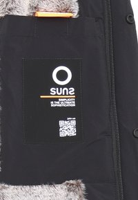 Giacca nera con colletto foderato in pelliccia e una toppa nera con lettere bianche, logo e codice QR. Include un dettaglio arancione sul colletto.