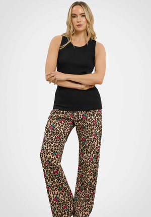 Donna in piedi con le braccia incrociate, indossa un top nero senza maniche e pantaloni con stampa leopardata e accenti floreali rossi e verdi.