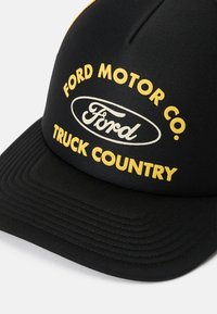 Černá čepice se strukturovaným deséningem, s žlutým textem, který říká "FORD MOTOR CO." a "TRUCK COUNTRY" nad bílým logem Ford. Texturovaný kšilt.