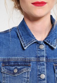 Farkkutakki, jossa on vaalea sininen pesu, klassinen kaulus, kaksi rintataskua, hopeiset napit ja erottuvat ompeluyksityiskohdat.