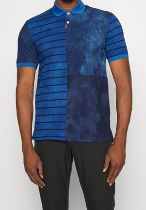 Man draagt een blauw poloshirt met korte mouwen met horizontale strepen aan de linkerkant en een gestructureerd marineblauw patch-ontwerp aan de rechterkant.