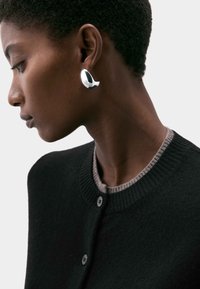 Stříbrné abstraktní náušnice na modelce, která nosí černý kardigan se šedým žebrovaným límcem a knoflíky. Vidět jsou detaily hladkého, lesklého povrchu.