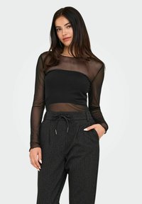 Kobieta z długimi ciemnymi włosami, ubrana w przezroczystą czarną bluzkę z długim rękawem na czarny biustonosz bandeau oraz spodnie w paski o wysokim stanie z elastycznym pasem.