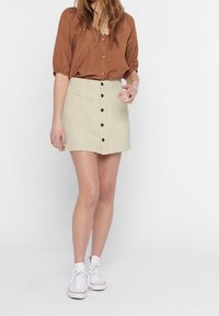 Gonna di cotone beige chiaro con vita aderente, dotata di bottoni davanti, due tasche frontali e un orlo leggermente svasato. Abbinata a una camicia marrone.