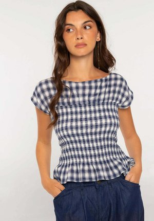 Mujer con cabello largo y oscuro que lleva una camiseta de manga corta a cuadros azul y blanca y pantalones azul oscuro con las manos en los bolsillos.