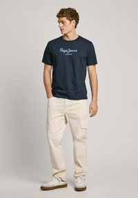 Tums zila kokvilnas T-kreklu ar balto "Pepe Jeans London" logotipu, apvienota ar gaiši beigu kargo biksēm un baltām sporta apaviem ar brūniem akcentiem.