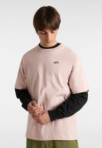 Ljusrosa bomulls T-shirt som är lager på lager över en svart långärmad skjorta; har en liten svart logotyp vid bröstet och en avslappnad passform.