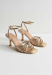 Sandali alti con tacco a spillo, open-toe, in oro, con un design frontale annodato, sottili cinturini alla caviglia e una finitura metallica liscia. Punta quadrata con un tacco moderato.