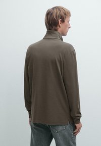 Braunes langärmliges Polo-Shirt mit einem hohen Kragen, weichem Material und seitlichen Schlitzen, getragen über hellgrauen Jeans.