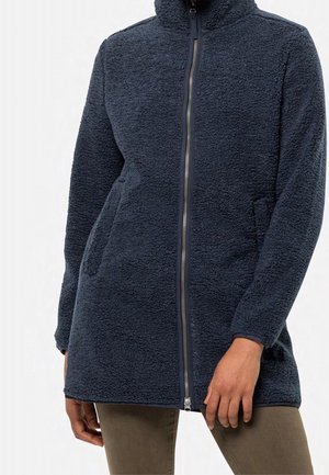 Mujer con chaqueta polar de color azul marino oscuro con cremallera frontal y bolsillos laterales, combinada con pantalones verde oliva.