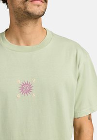 Lichtgroene katoenen t-shirt met een centraal zonontwerp in paars en crème, met de letters "RVCA" eromheen. Korte mouwen en ronde hals.