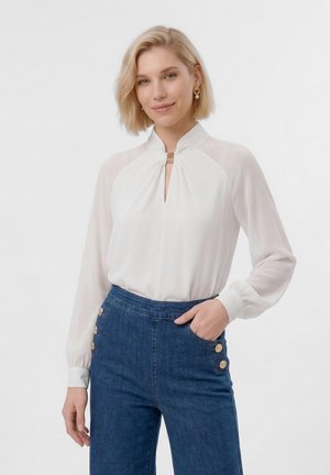 Blondína v bielej blúzke s dlhými rukávmi a vysokým golierom, oblečená v modrých džínsoch s vysokým pásom a zlatými gombíkmi, mierne sa usmieva.