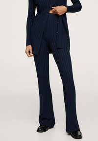 Osoba oblečená v námořnicky modrých ribkovaných pletených kalhotách a odpovídajícím dlouhém kardiganu, doplněná černými koženými mokasíny, stojící na neutrálním pozadí.