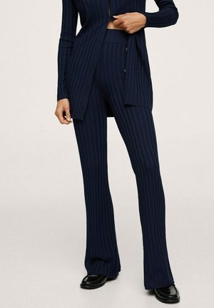 Persoon in marineblauwe geribbelde gebreide broek en bijpassende lange cardigan, gecombineerd met zwarte leren instappers, staand tegen een neutrale achtergrond.