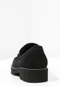 Svart mockaskor med rund tå, med en texturerad yta och en tjock sula med glittrande accenter. Bakifrån framhäver bilden skons eleganta design.