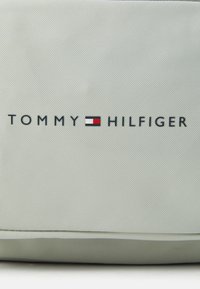 Ljusgrå väska med en texturerad yta, som pryds av "TOMMY HILFIGER"-logotypen i svart, accentuerad av ett färgat rektangulärt flagg-emblem.