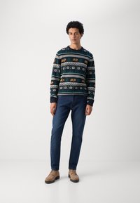 Gehaakte trui in donkerblauw met kleurrijke geometrische patronen, geribbelde mouwen en zoom; gecombineerd met donkere jeans en lichtbruine schoenen.