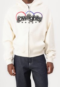 Felpa zip-up color crema con una grande grafica nera "lowlights", contorni curvi rossi e blu, e una tasca a marsupio. Presenta polsini e orlo a costine.