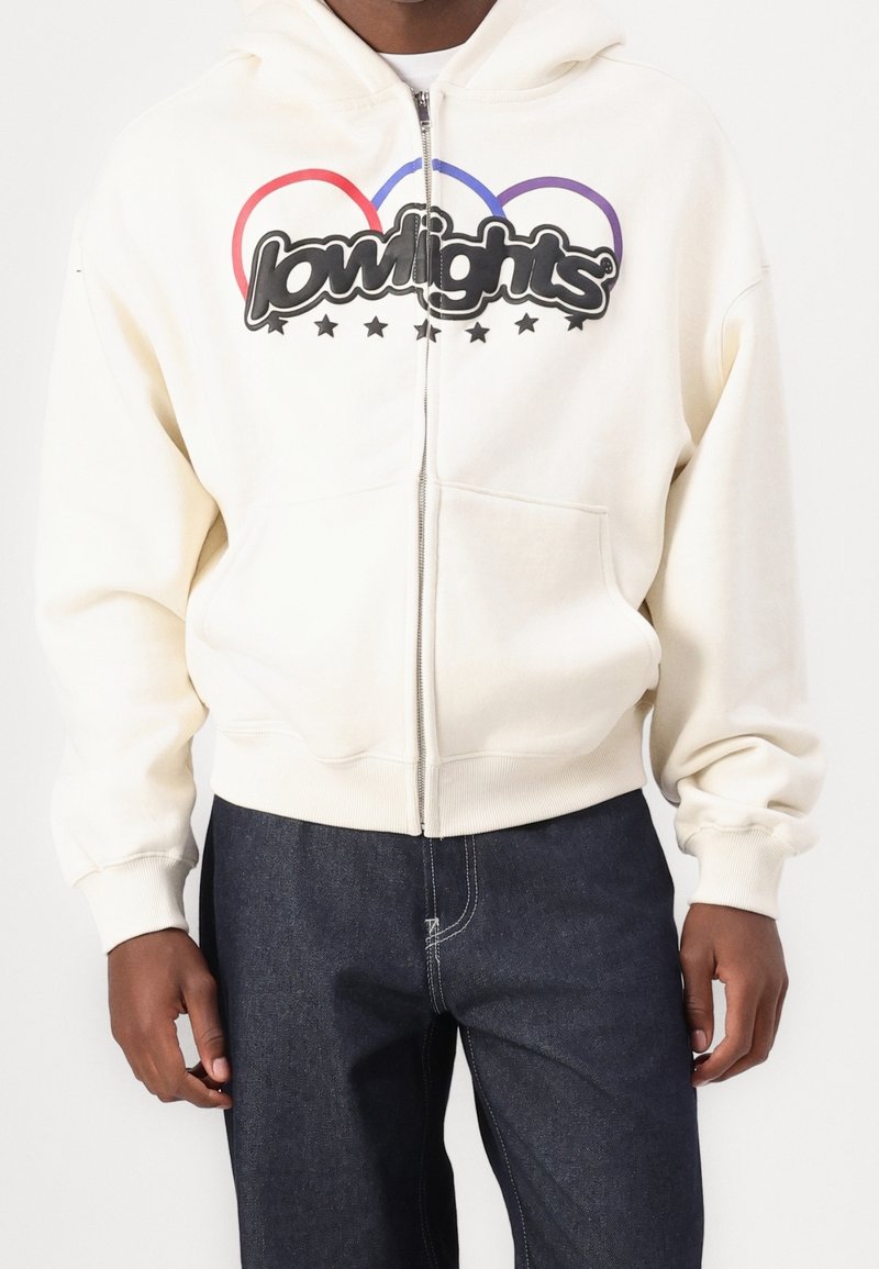 Felpa zip-up color crema con una grande grafica nera "lowlights", contorni curvi rossi e blu, e una tasca a marsupio. Presenta polsini e orlo a costine.