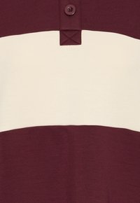 Camisa a rayas burdeos y crema con tapeta de botones y un parche texturizado debajo de la raya superior. Tejido suave, diseño casual.