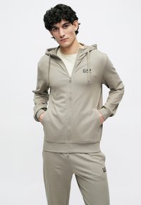 Fiatal férfi göndör hajjal, világos bézs színű, cipzáras EA7 logós kapucnis pulóvert és hozzá illő nadrágot visel, zsebeiben tartja a kezét.