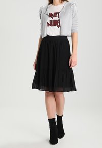 Grå kort jacka över en vit t-shirt med röd text, kombinerat med en svart plisserad kjol. Modell bär svarta ankelskor.