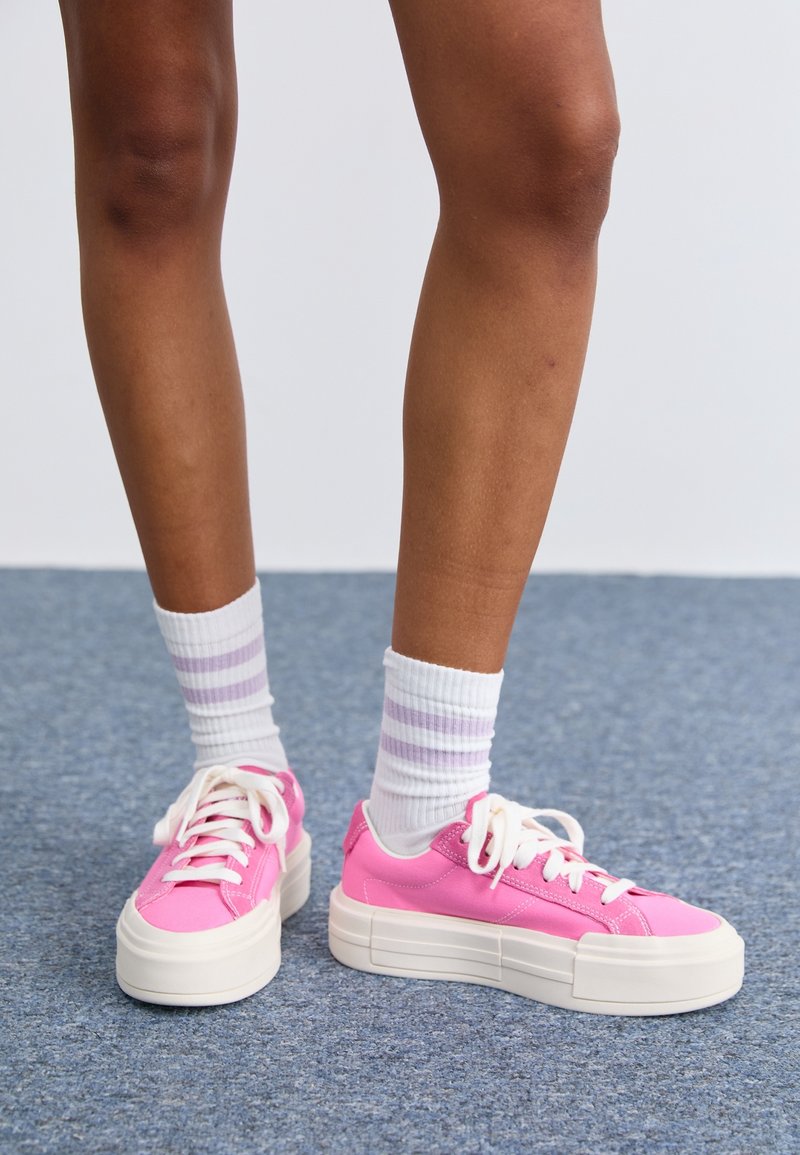 Des baskets roses avec une semelle blanche épaisse, dotées d'un design à lacets et d'un matériau supérieur lisse. Portées avec des chaussettes blanches munies de rayures violettes.