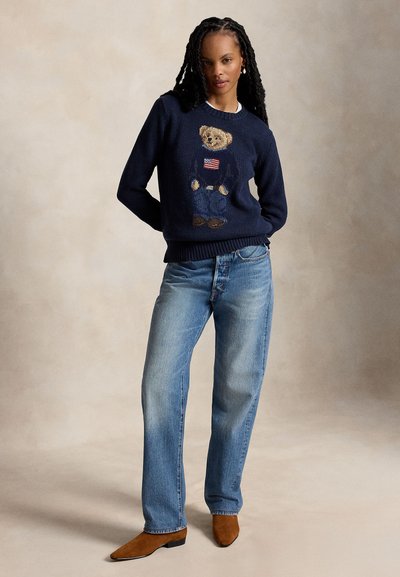 Moteris dėvi tamsiai mėlyną megztinį su meškiuku, laikantį Amerikos vėliavą, ir mėlynas džinsus, stovinti rankomis už nugaros neutraliame fone.