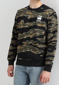Kamouflagesweatshirt i svart, grön och beige. Har rundad halsringning, ribbade ärmslut och en vit logotyp på bröstet. Tillverkad av bomullsblandning.