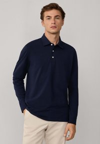 Granatowa koszula polo z długim rękawem i kołnierzykiem oraz zapięciem na trzy guziki, wykonana z miękkiego materiału, w zestawieniu z jasnymi spodniami.