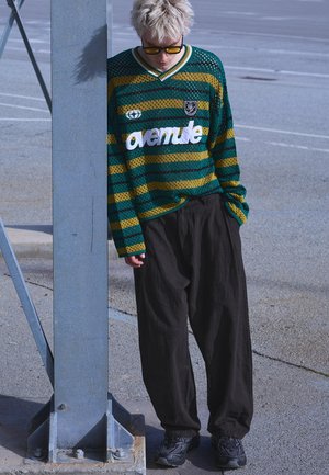 Young person with short bleached hair wearing yellow-tinted glasses, green and yellow striped sweater with "avenue" text, and loose black pants.