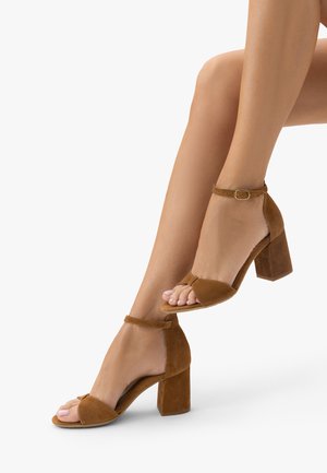 Nogi kobiety w brązowych zamszowych sandałach na blokowym obcasie z odkrytymi palcami i paskami wokół kostki na białym tle.