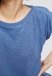 Modrý texturovaný top s širokým výstřihem a nařasením na ramenech, vyrobený z měkké látky vhodné pro každodenní nošení.