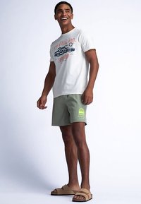 Světlé šedé tričko s motivem auta, zelené plavky s žlutým logem a hnědé sandály, modelované usměvavou postavou.