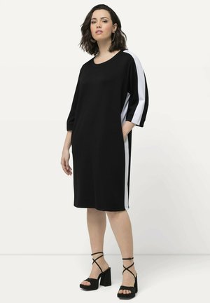 Zwarte knie-lange jurk met driekwart mouwen en witte zijpanelen; gladde textuur; gestyled met zwarte plateausandalen.