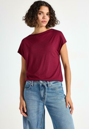 Kvinde med krøllet hår iført en burgunder kortærmet top og lyseblå højtaljede jeans med en bundet bælt, der står foran en hvid baggrund.