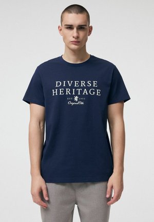 Młody mężczyzna w granatowym t-shircie z napisem "Różnorodne dziedzictwo" i szarych spodniach, stojący na jednolitym jasnym tle.