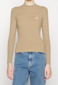 Haut beige tricoté à manches longues avec un col montant et une texture côtelée, orné d'un petit logo brodé sur la poitrine, associé à un jean bleu.