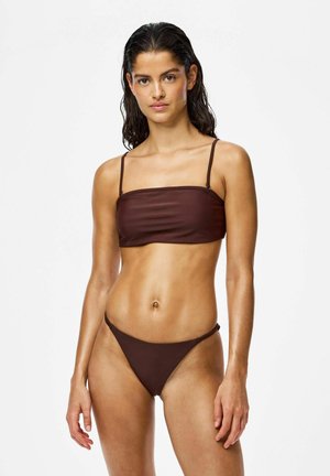 Žena s mokrými vlasmi, oblečená v tmavohnedom dvojdielnom bikinách s tenkými ramienkami, stojaca pred jednofarebným svetlým pozadím.