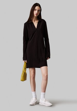 Zwarte lange jurk met een kraag en een V-hals. Gepaard met een gele tas, witte sneakers en gestructureerde witte sokken.