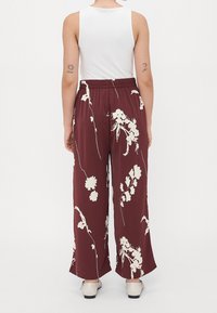 Personne portant un pantalon large bordeaux avec un imprimé floral blanc, un haut blanc sans manches et des chaussures crème, vue de dos.