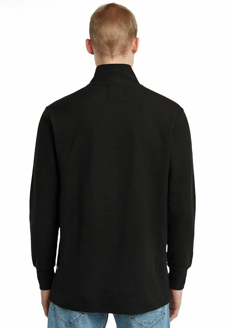 Schwarzes Langarmshirt mit geradem Schnitt, weichem Material, Kragen und seitlichen Schlitzen; getragen über hellblauen Jeans.