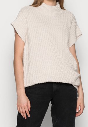 Vrouw draagt een crèmekleurige gebreide trui met korte mouwen en een hoge hals, met een zwarte broek, zichtbaar van schouders tot heupen.