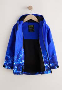 Veste imperméable bleue avec motif d'éclairs sur les manches et l'ourlet, doublure intérieure noire, suspendue ouverte sur un cintre en bois sur fond blanc.