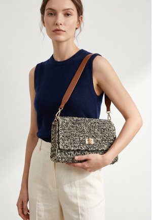 Femme portant un haut sans manches bleu marine et un pantalon crème tient un sac à main tissé noir et blanc avec une bandoulière marron sur l’épaule.