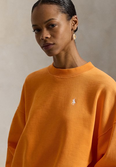 Πορτοκαλί φούτερ με ριμπ λαιμόκοψη, άνετη εφαρμογή και κεντητό λευκό λογότυπο. Υφασμάτινη υφή, εμφανείς ραφές.